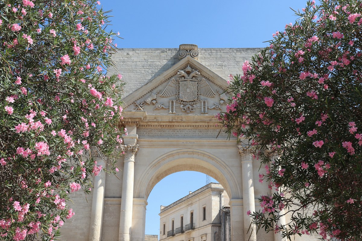 A carved stone city gate framed by pink oleander in Lecce